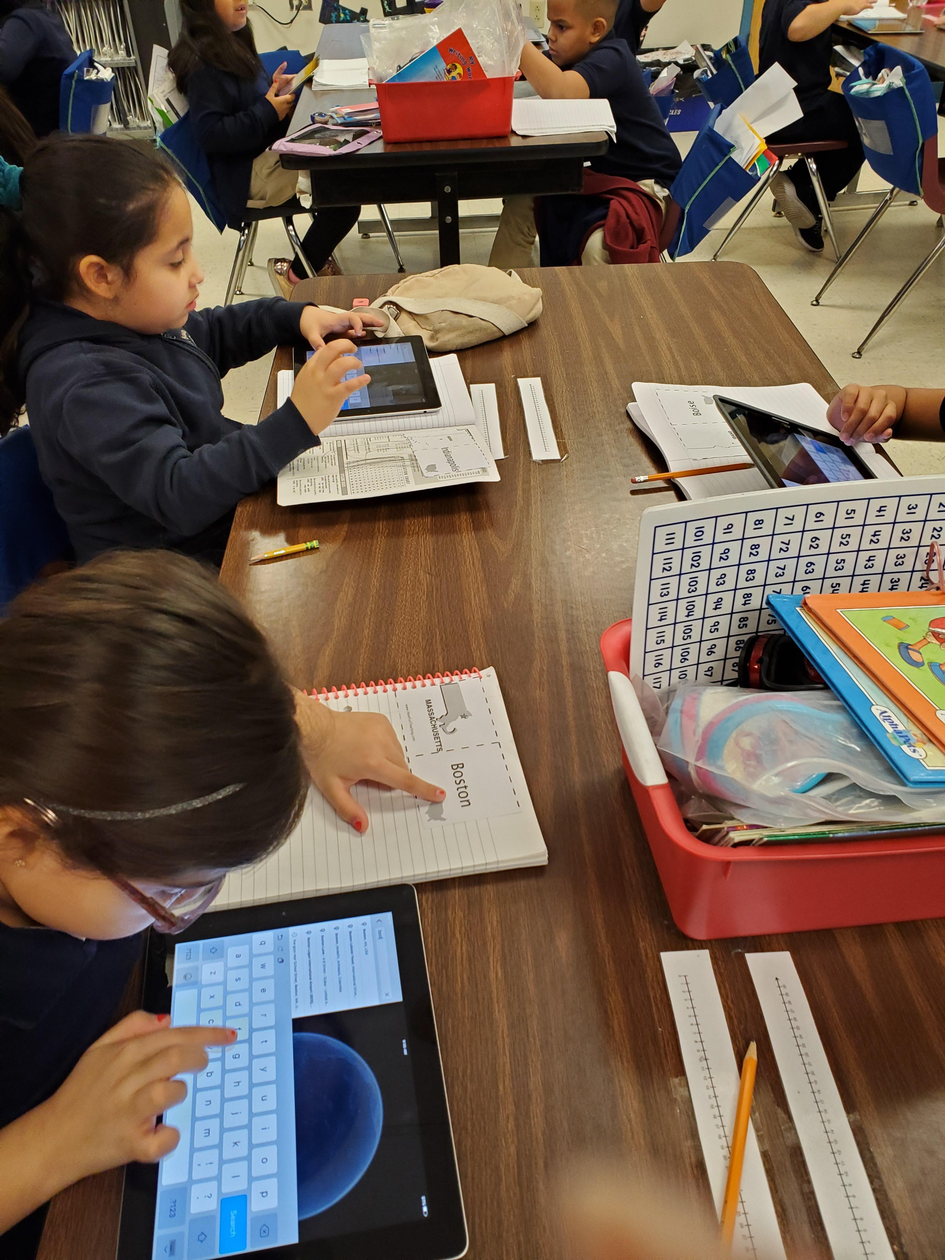several children seated at desks with designated cities and using ipads to look the up