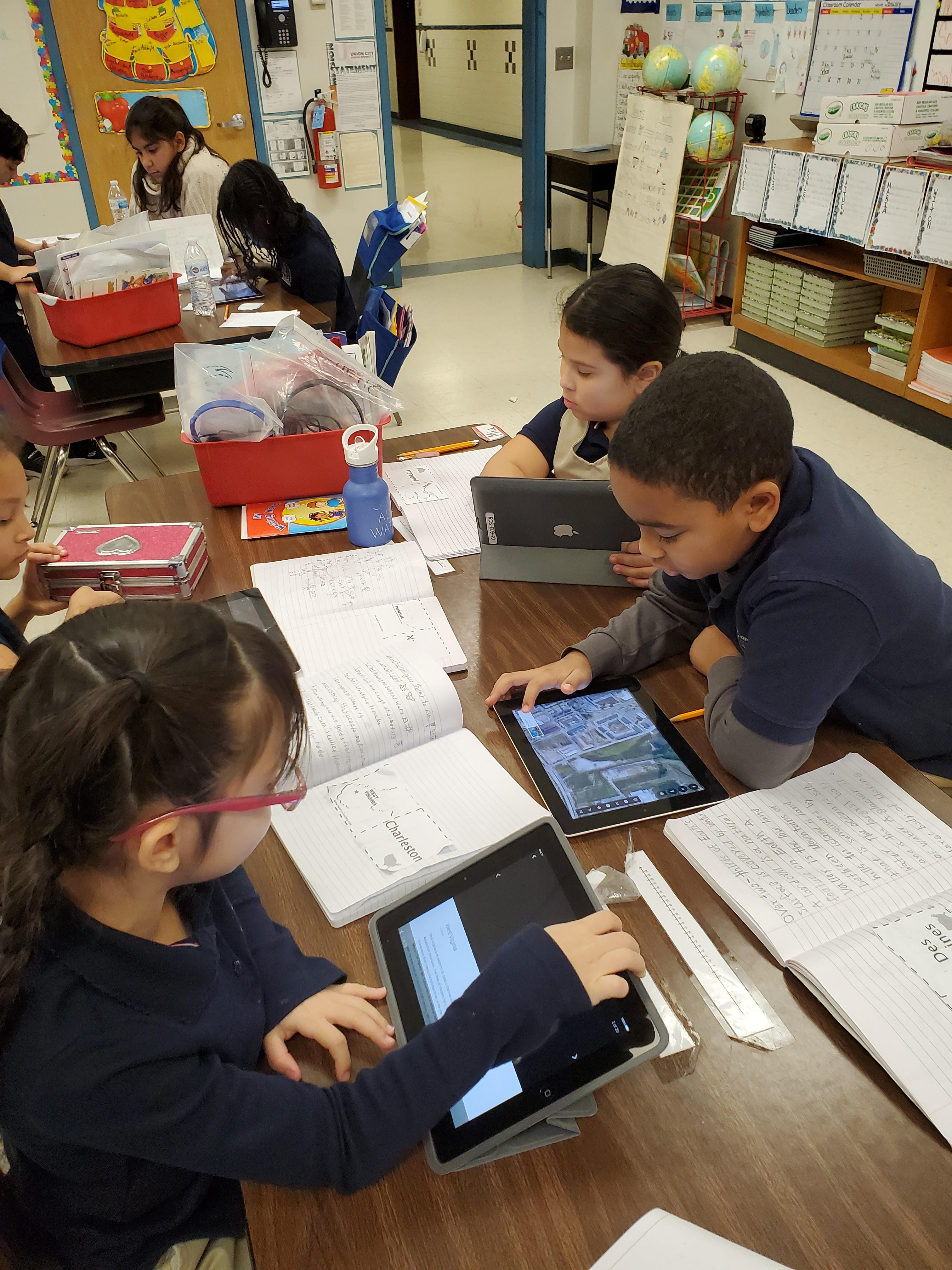 several children seated at desks with designated cities and using ipads to look the up